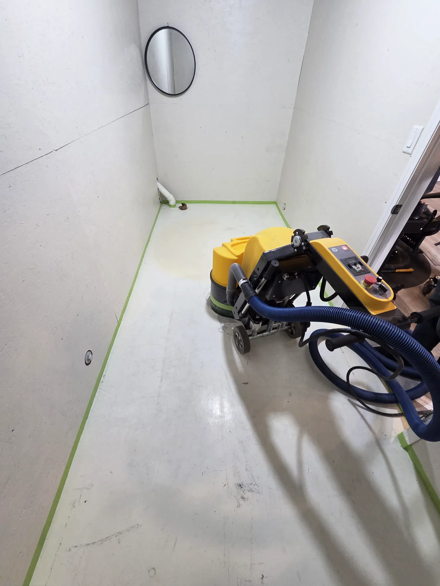Yellow walk-behind concrete grinder with a dust-extraction hose preparing a floor, edges masked with green tape