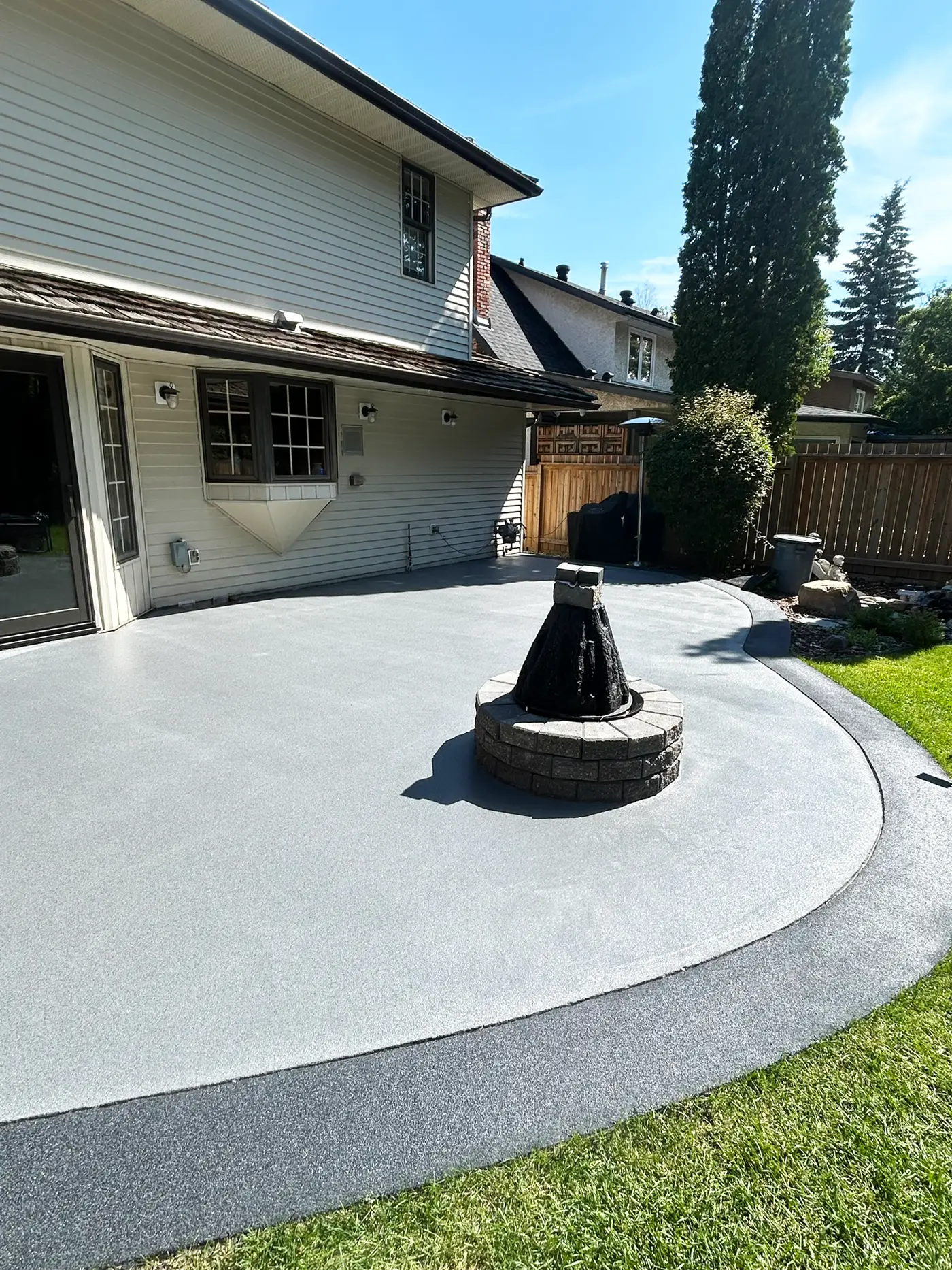 Grey rubber pavement patio with a darker border curving around a stone fire pit behind a two-storey home