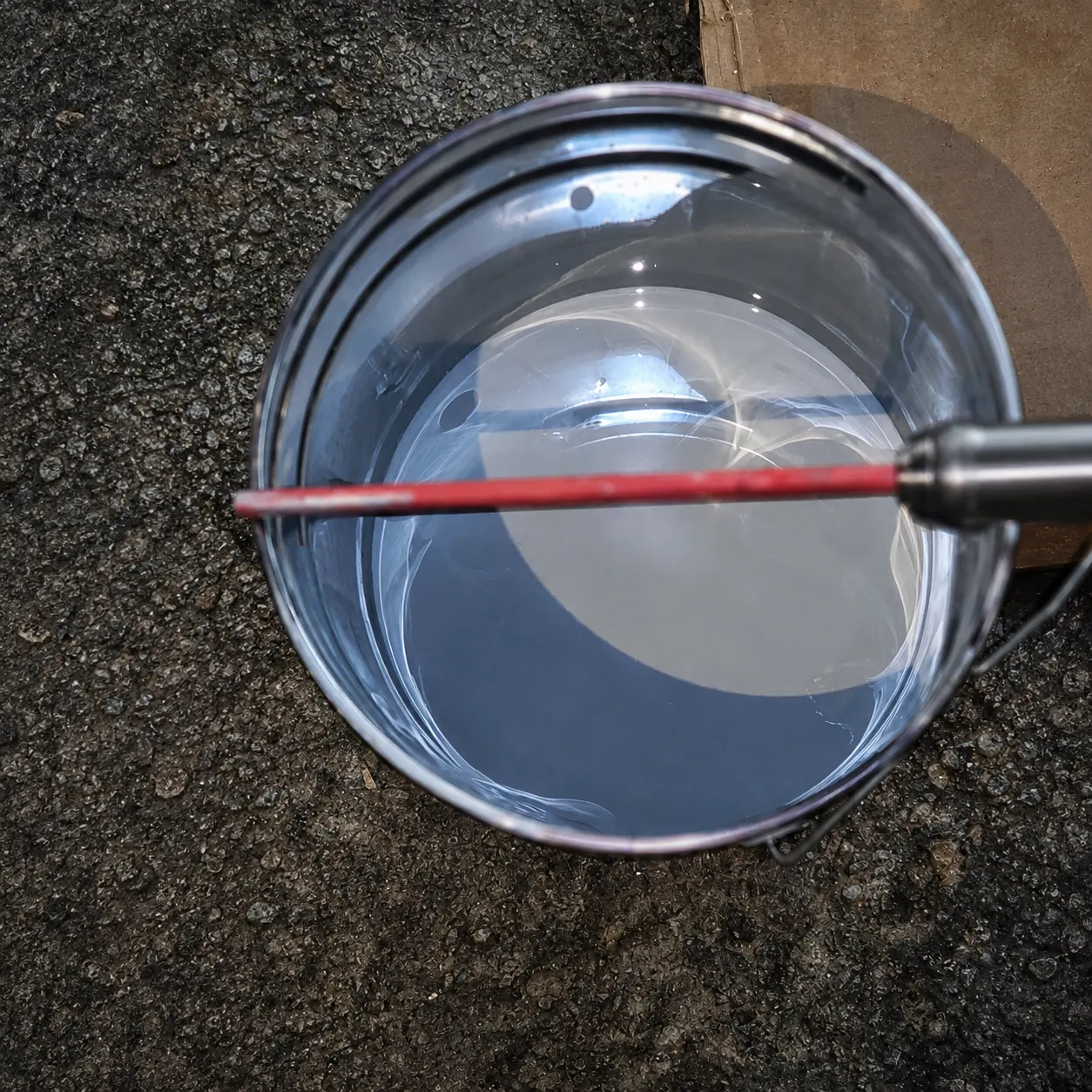 Steel pail of mixed clear coating resin with a red mixing paddle resting on a dark aggregate surface